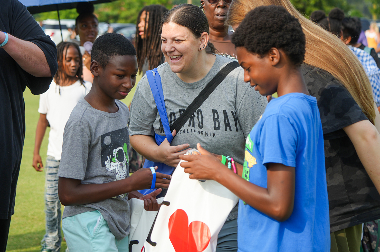 A woman holding a bag hands out items to two children, one wearing a blue shirt, the other gray.