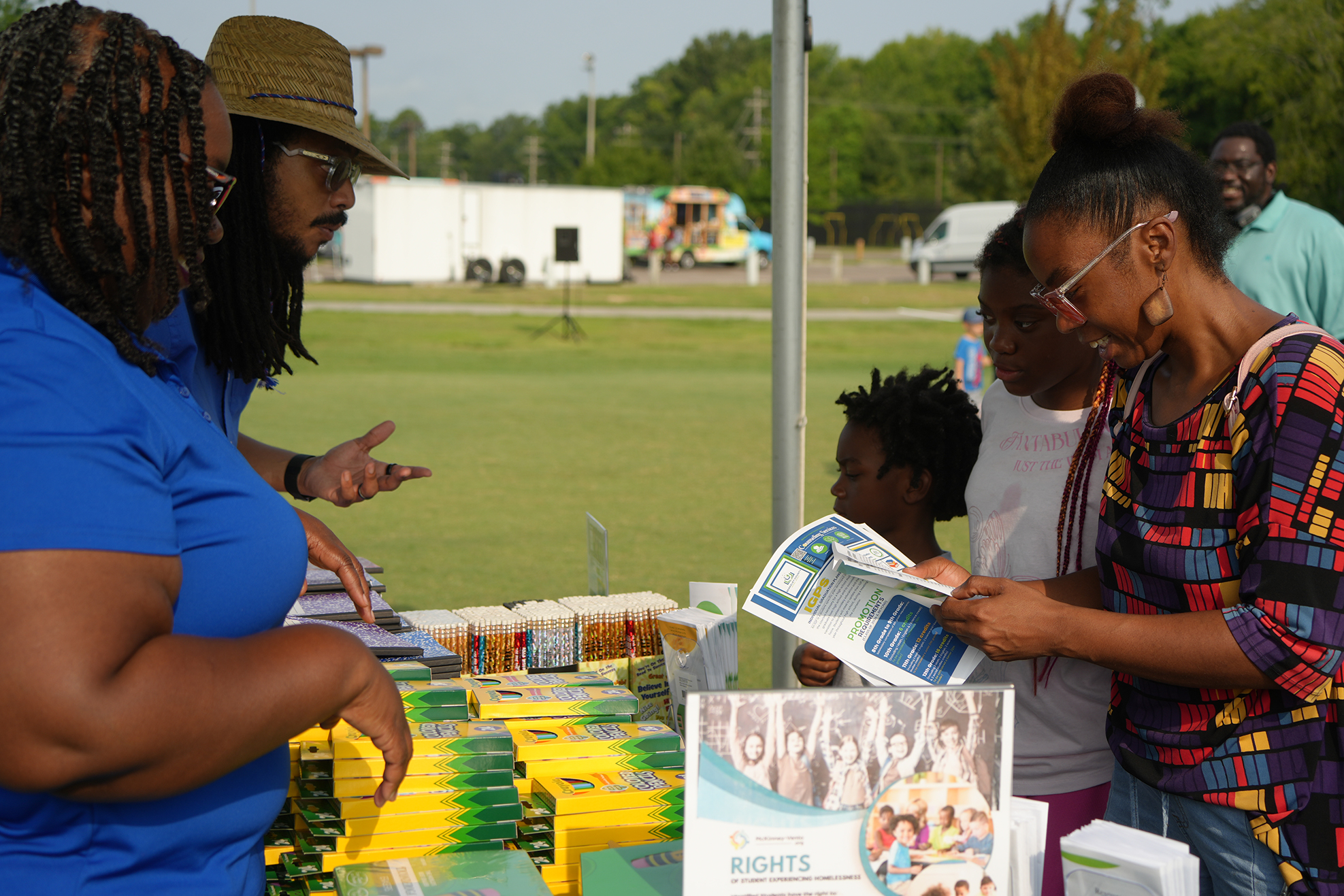 People under a canopy view pamphlets on a table with yellow and green boxes; a grassy field is in the background.