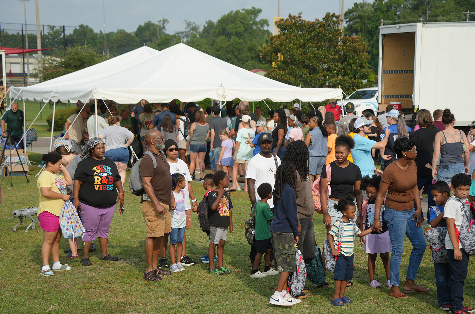 Numerous people gathered under a large white tent. Some are standing, some are walking. Several wear sunglasses. A white box truck is parked in the background.