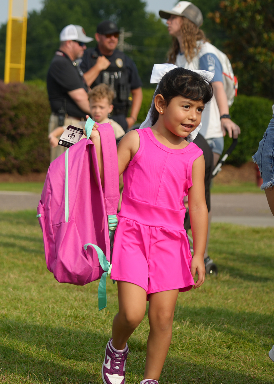 A young girl in a pink dress walks in a park, holding a pink backpack with green straps.