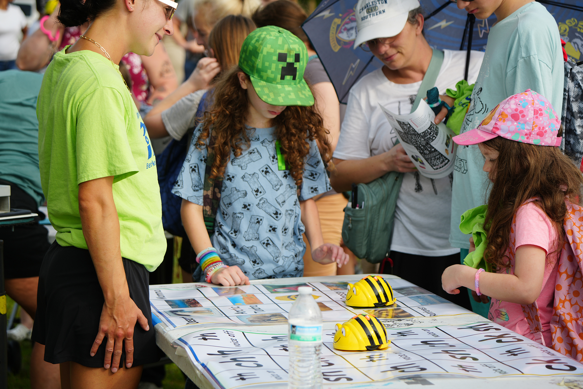 A woman and two girls examine a board at a table. The board contains a map and bee cutouts.