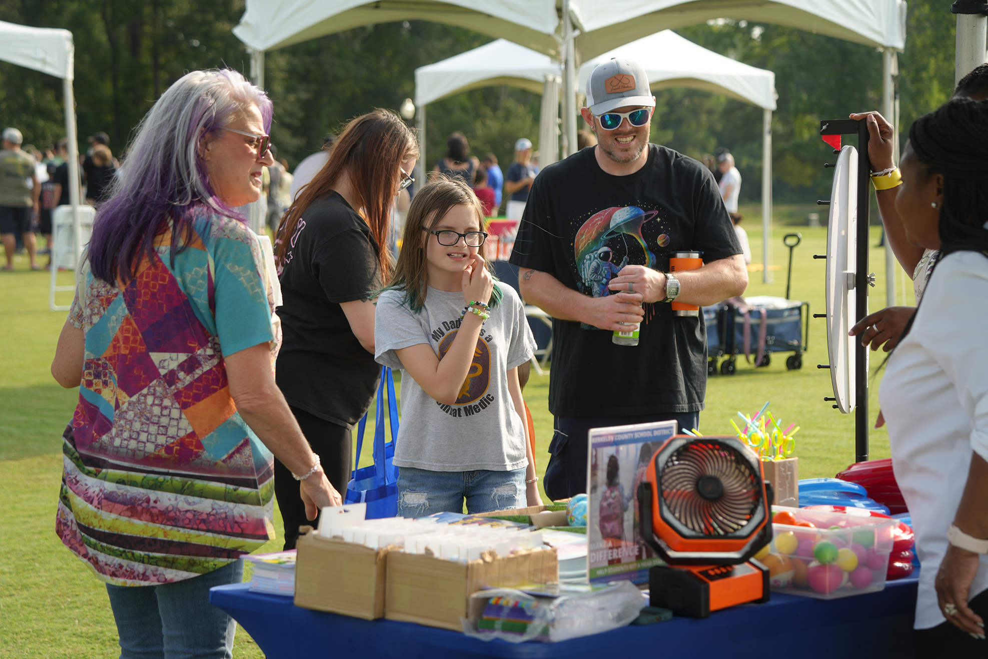 A group of people gather around a table with items on it, including a fan and other goods.
