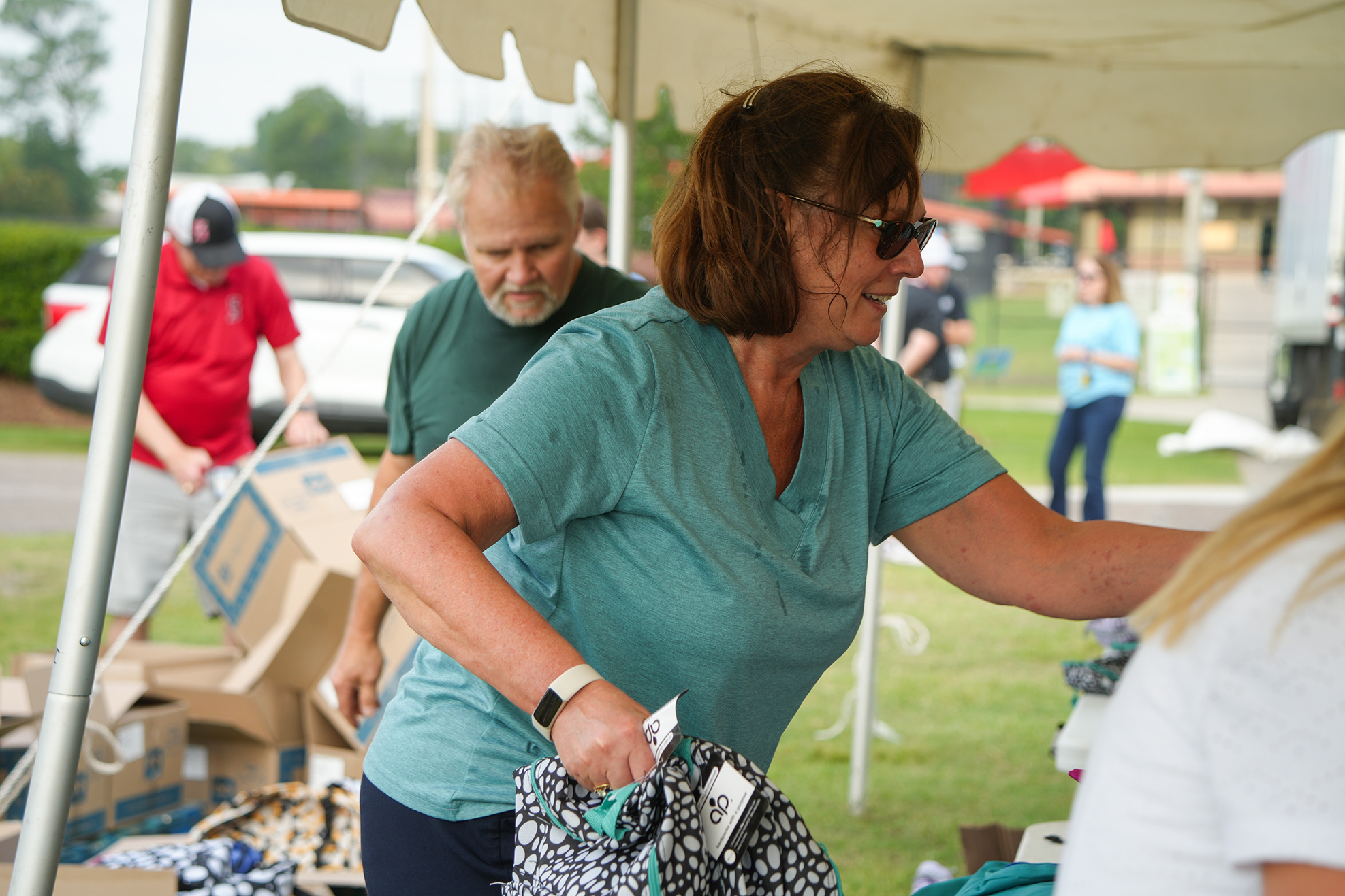 A woman in a teal shirt holds a bag under a tent, surrounded by boxes and people.