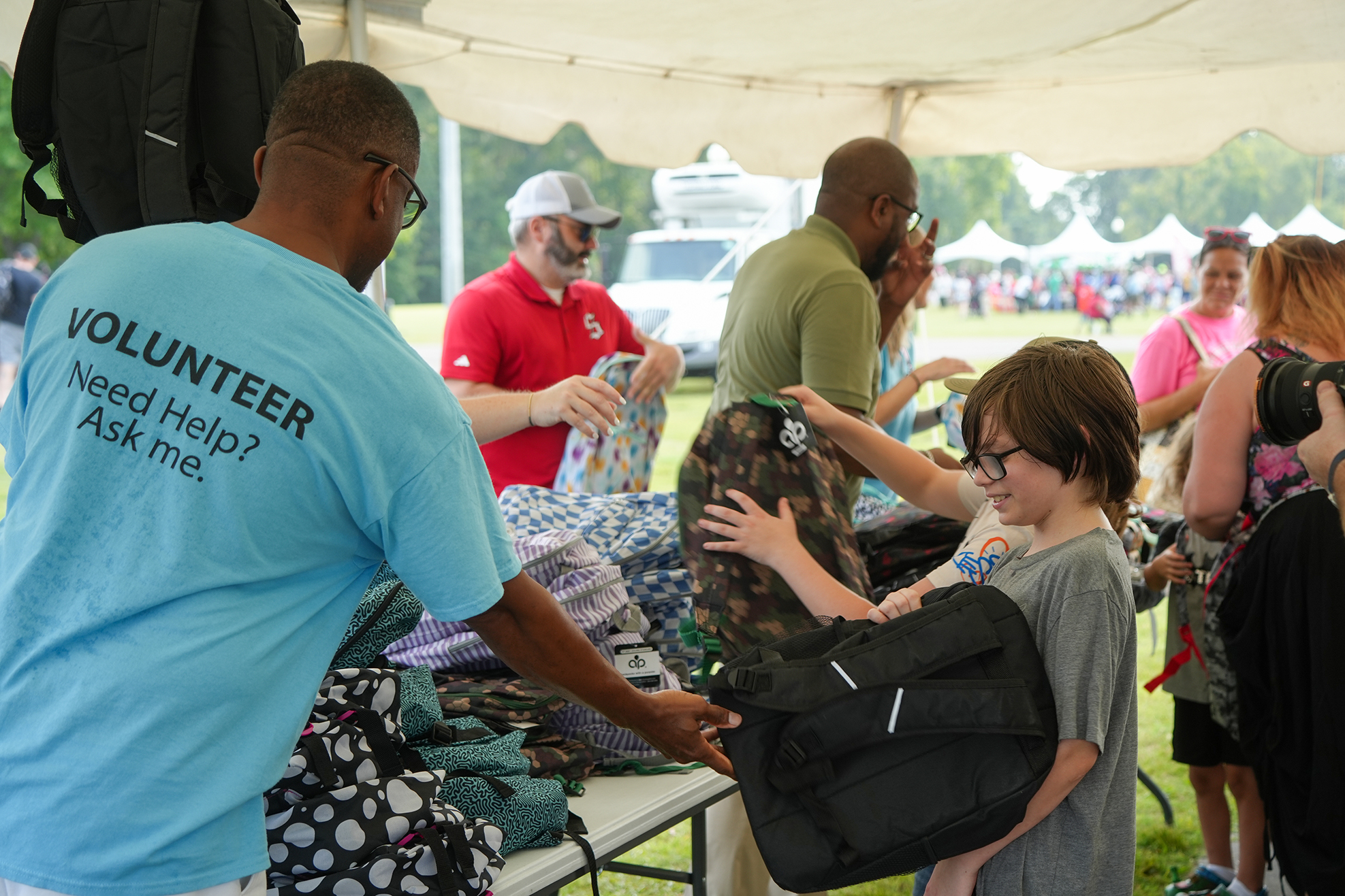 A man in a blue shirt helps a boy pick out a black bag at a community event.