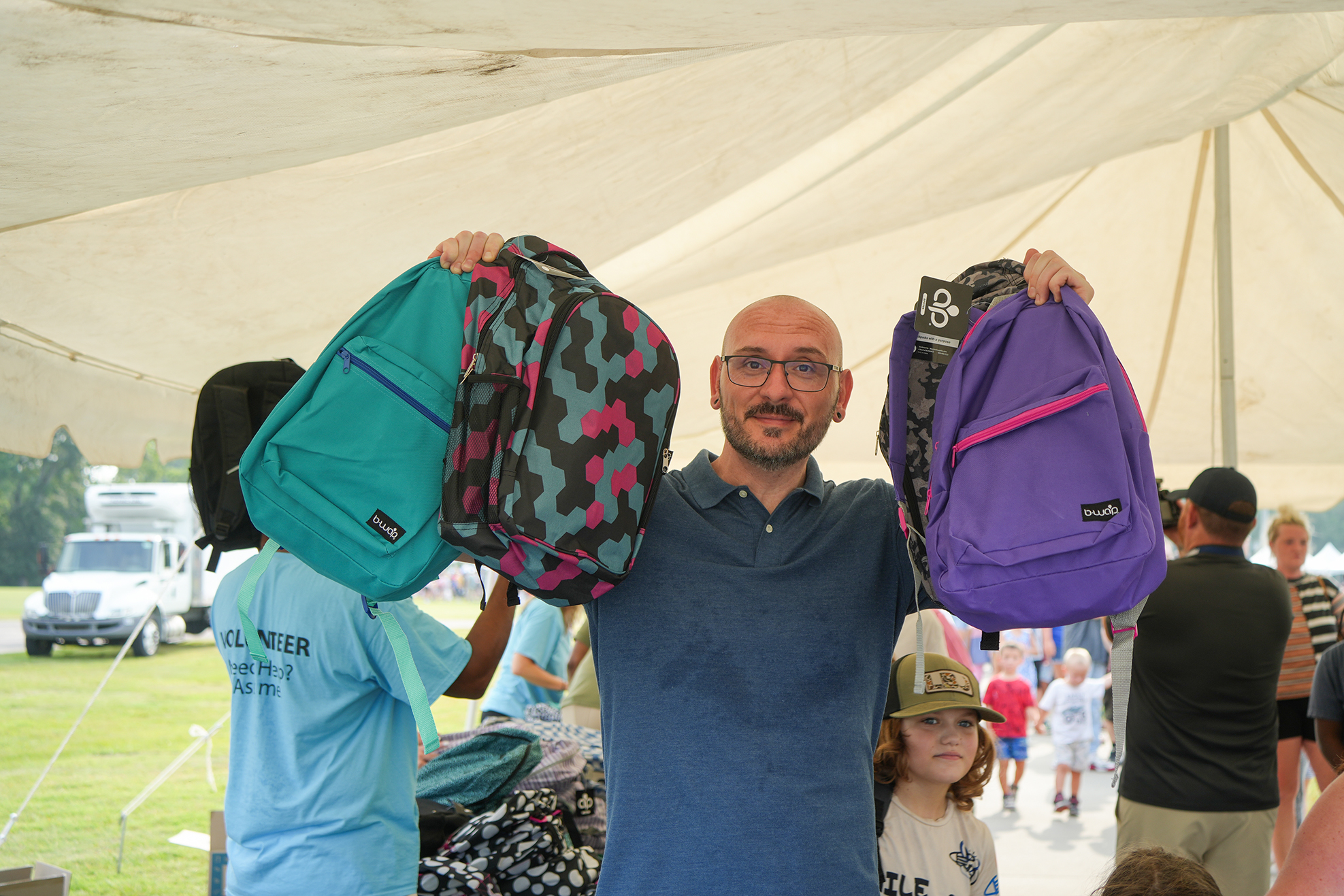 A man holds two backpacks, one teal and one purple, under a tent. People in the background include a child and adults.