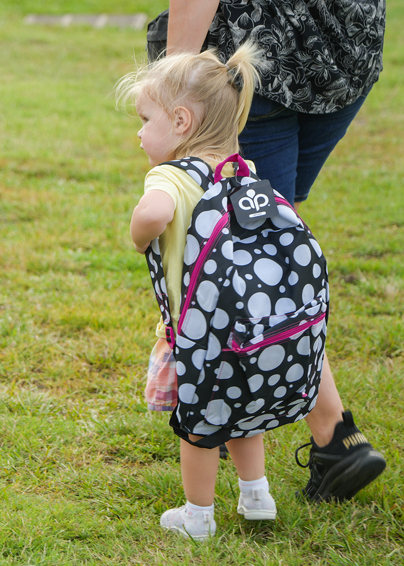 A toddler, wearing a backpack, stands on a grassy field, facing away from the camera. A person wearing blue jeans walks behind the toddler.