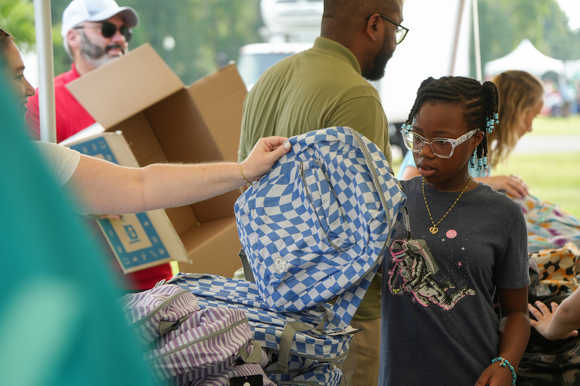 A girl with glasses holds a backpack, while an adult hands her another bag. A tent is in the background.