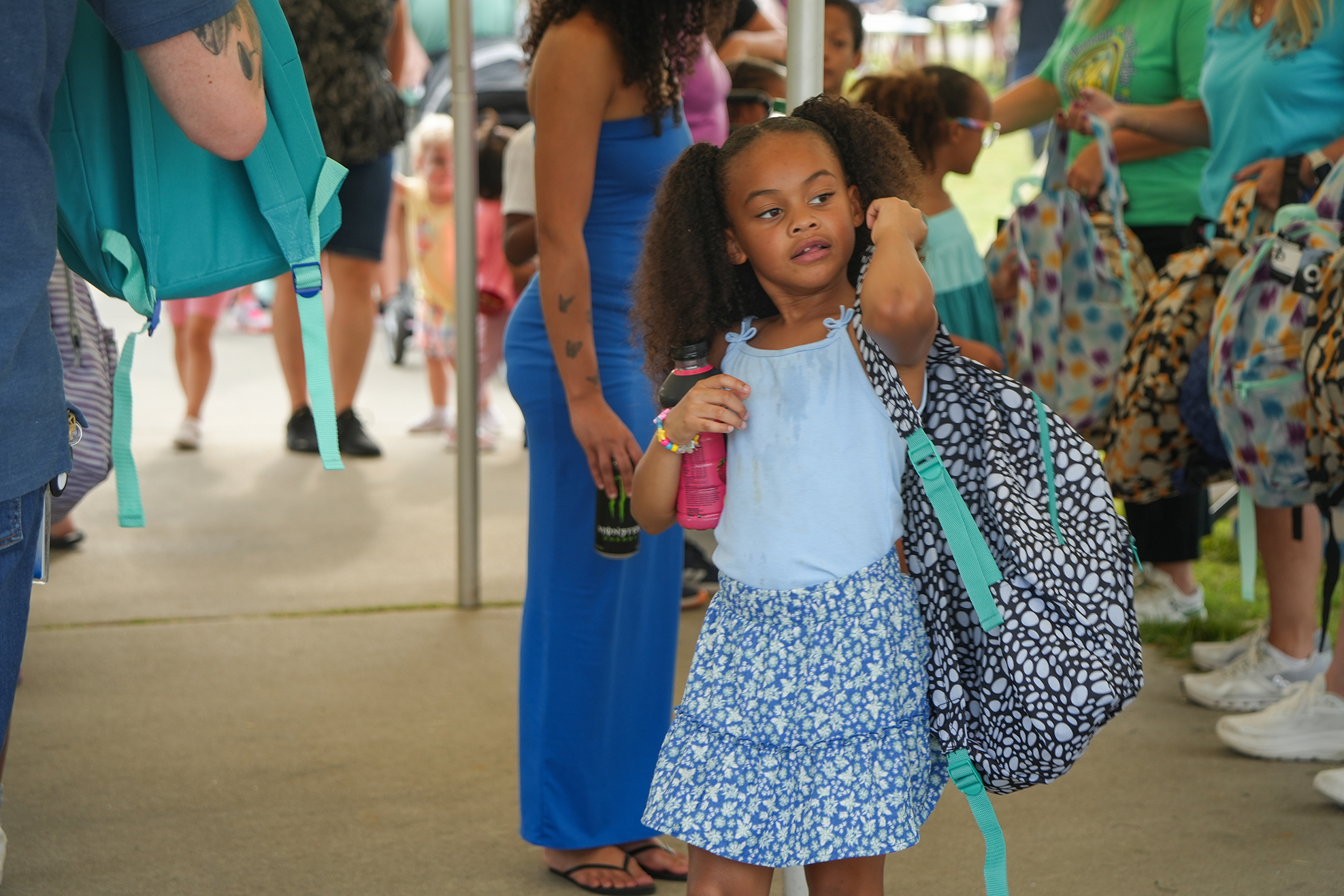 A young girl with a blue skirt and floral top walks holding a pink bottle and backpack.