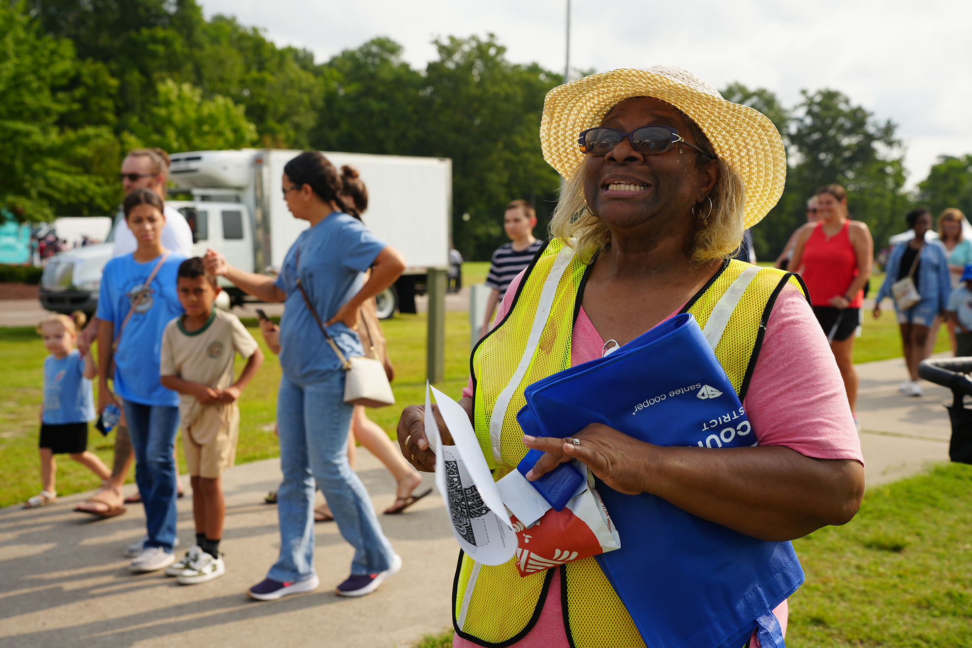 A woman in a yellow vest holds a blue folder. People in the background walk in a park.