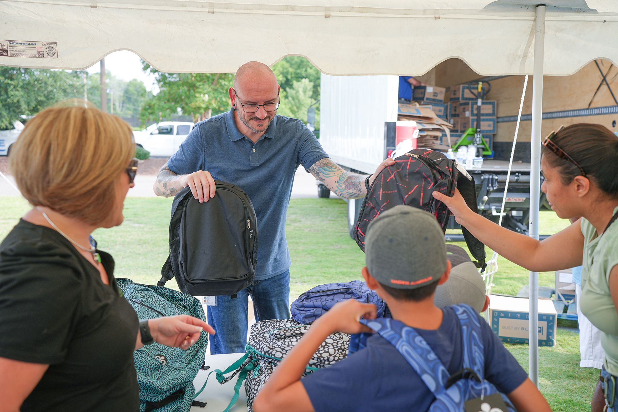 A man with tattoos hands a backpack to a woman and another to a child under a tent.