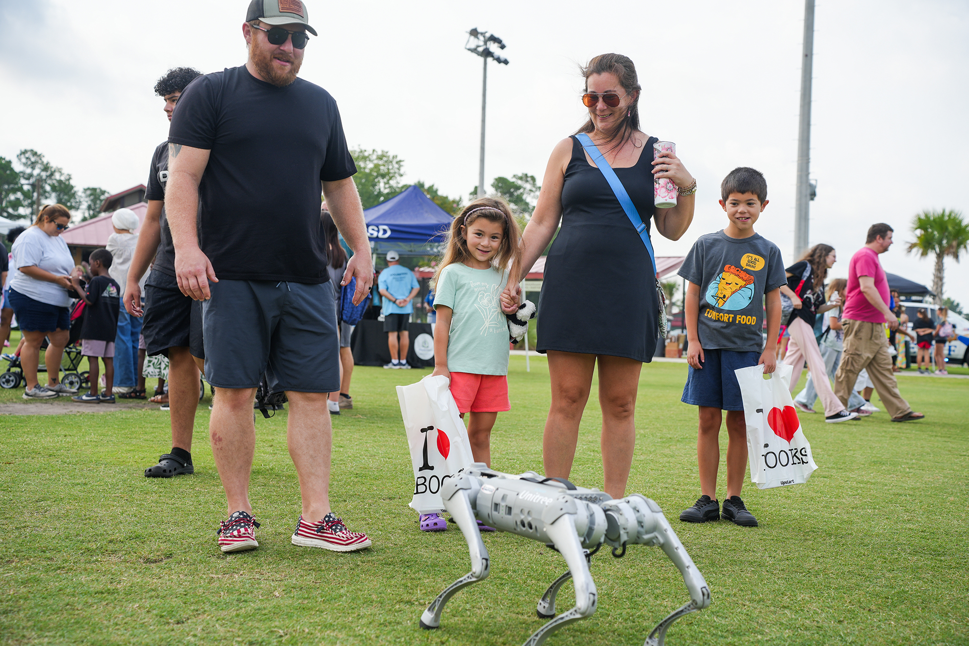 Family with two kids next to a robotic dog. Behind them, several people walking. Trees and canopies in the background.