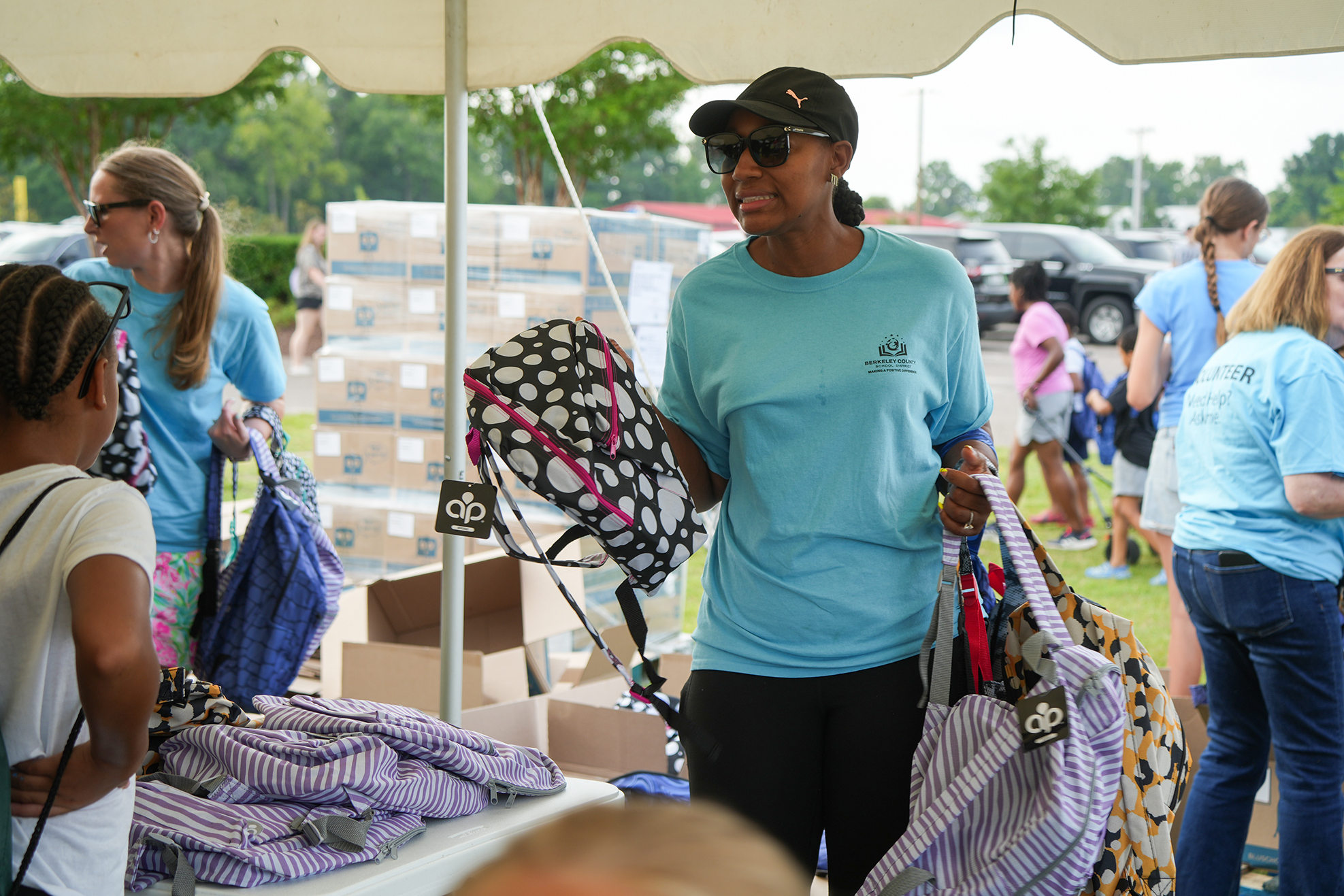 Person in light blue shirt and cap holds bags near table with supplies. Many people in light blue shirts.