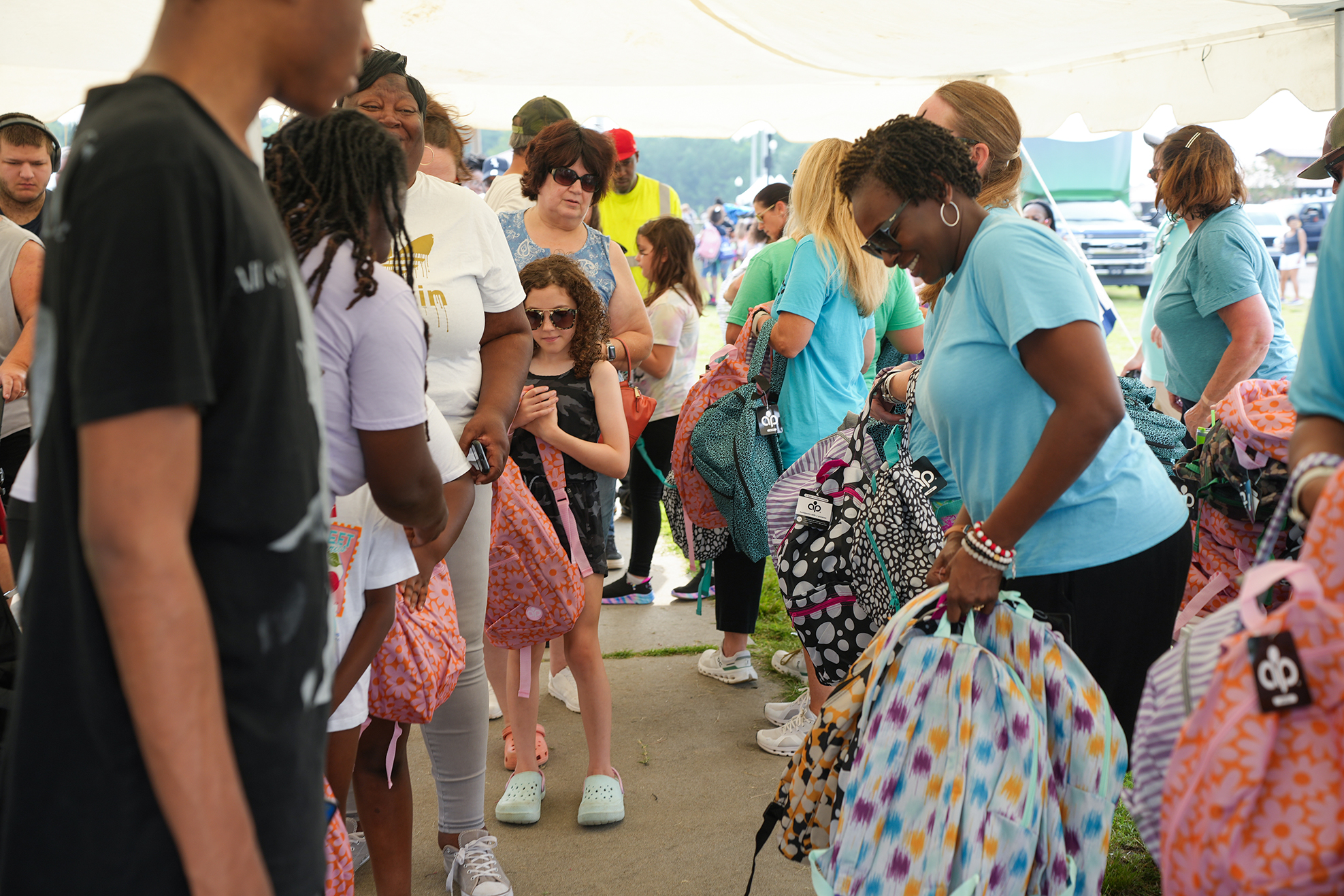A group of adults and children are in a tent. Some hold colorful backpacks, others look at their phones.