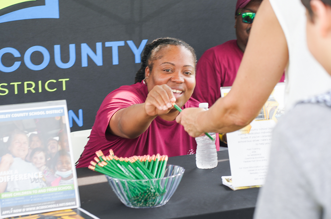 A woman sits at a table with green pencils, a water bottle, and brochures. She hands a pencil to a person.