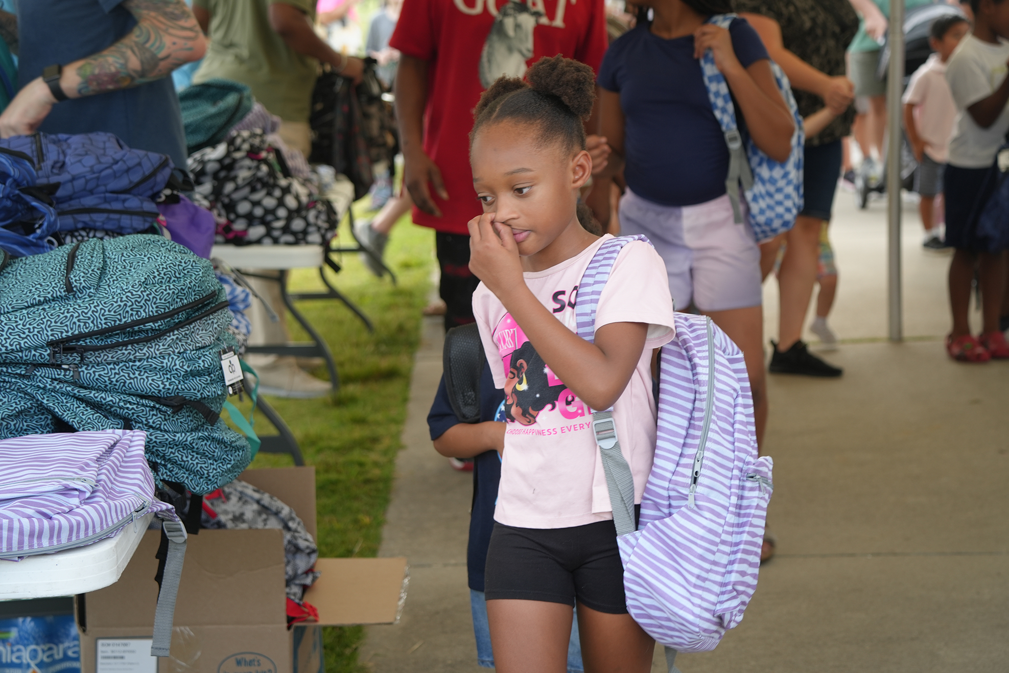 A girl with a backpack walks past a table with bags and boxes. People stand and walk in the background.