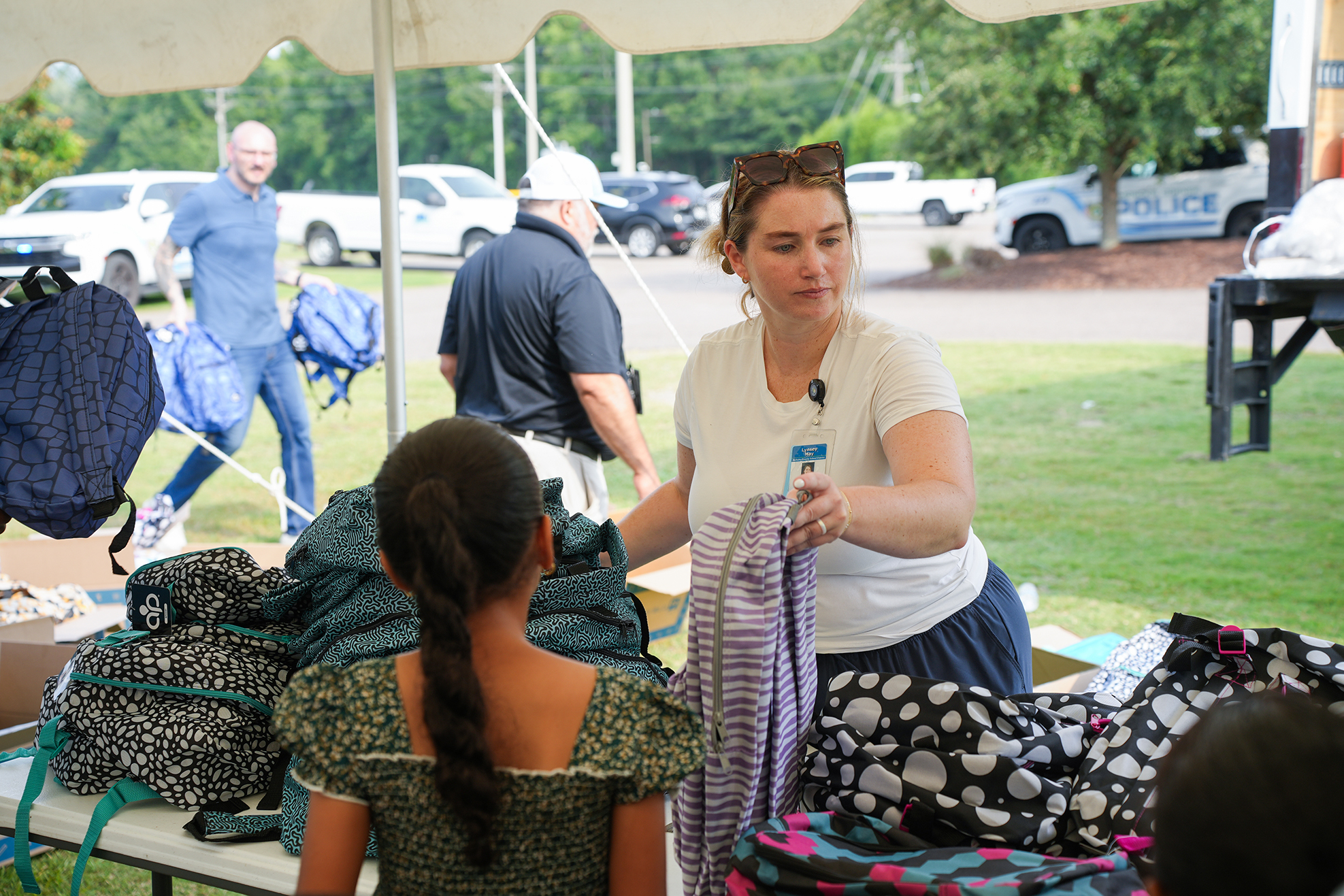 Woman in white shirt helping girl with ponytail select backpacks. Various bags and backpacks are on a table.