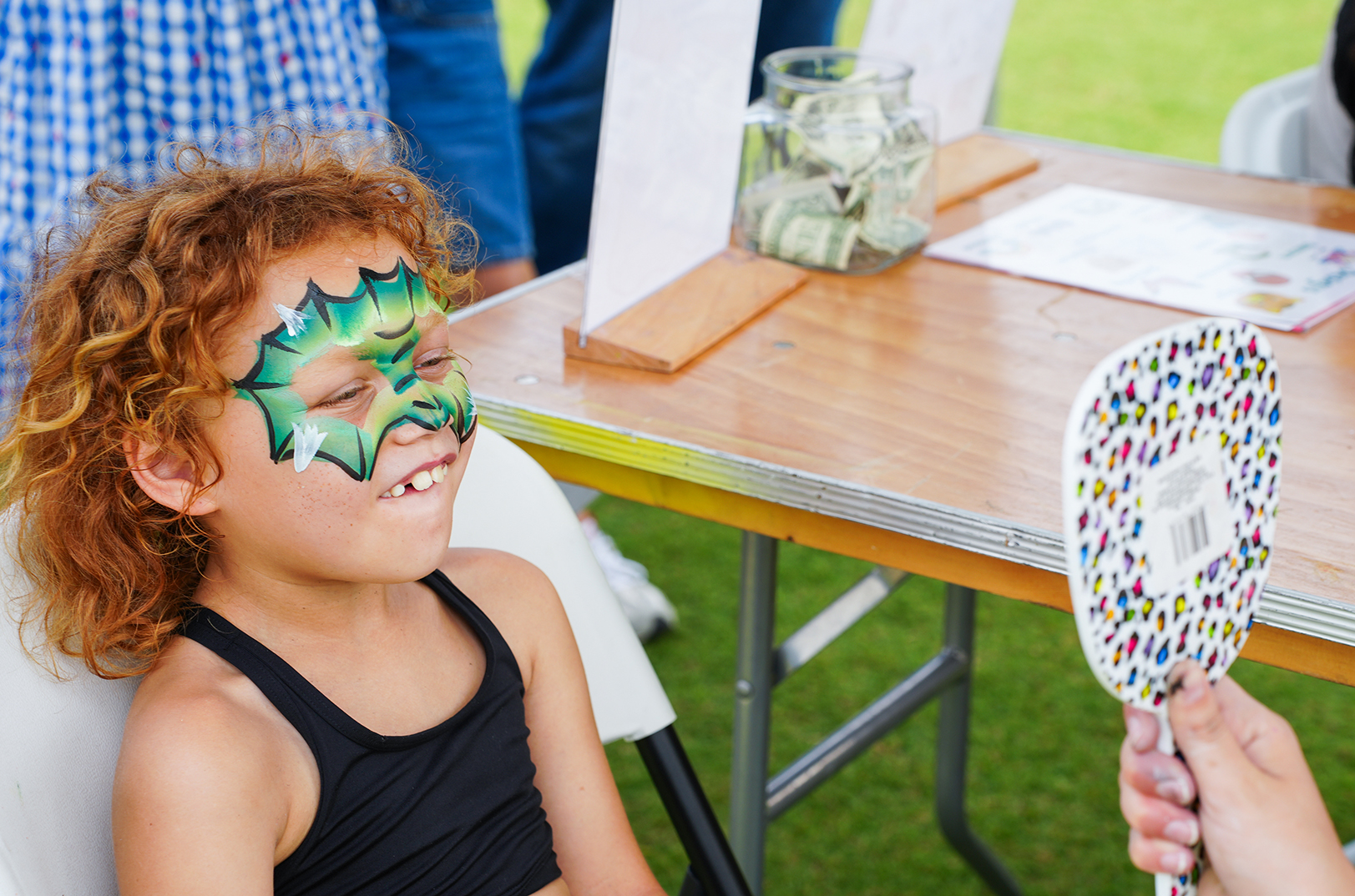 A child with red curly hair is seated, smiling, wearing a green mask with leaf patterns. A person holds a colorful fan.