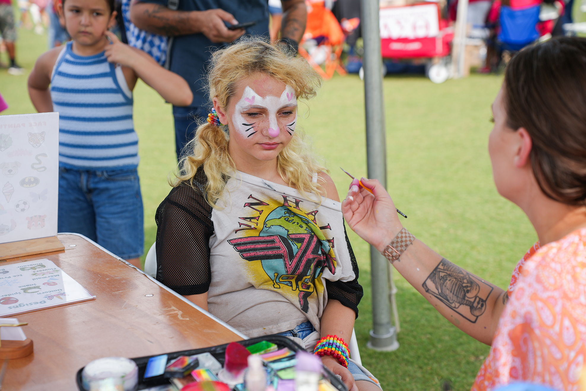 A woman paints a face design on a young girl, who sits under a tent at an outdoor event.