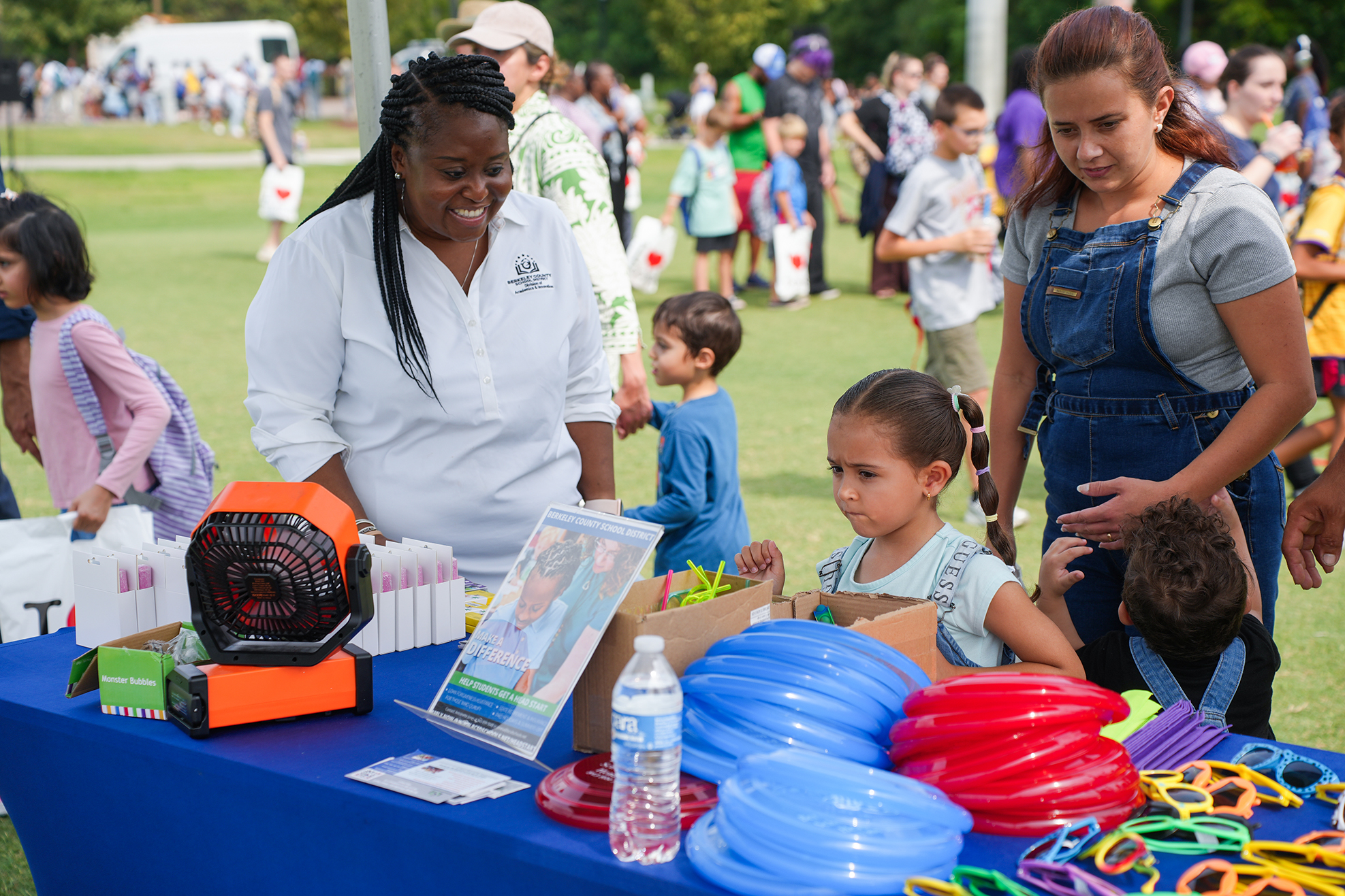 Two women and a girl are at an outdoor event, with toys and a table. Other people are in the background.