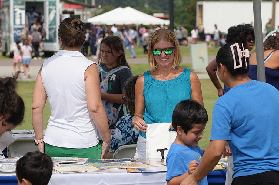 Adults and children gather around a table with books. One woman wears sunglasses. In the background, people walk past trucks.