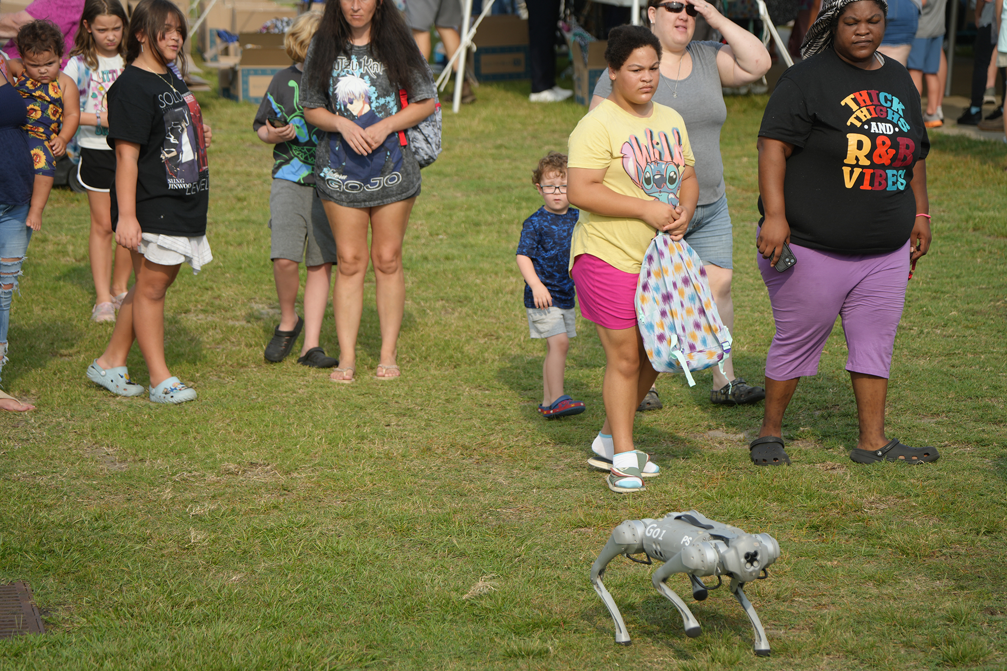 People stand on grass, some watching a small, mechanical dog. A person in yellow and pink shorts holds a blanket.