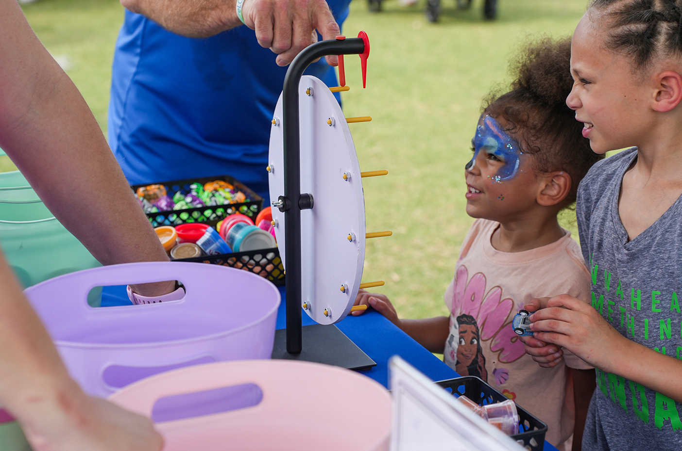 Two children and an adult at a table with a spinner and colorful face paint on a sunny day.