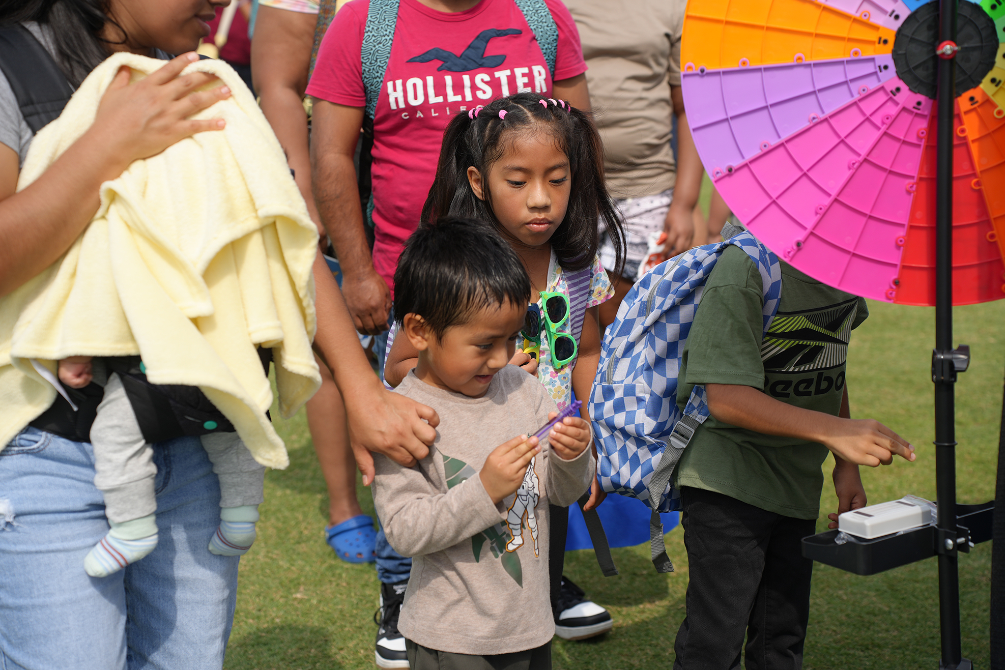 Several children gather around a colorful wheel. One child holds a necklace and another holds sunglasses.