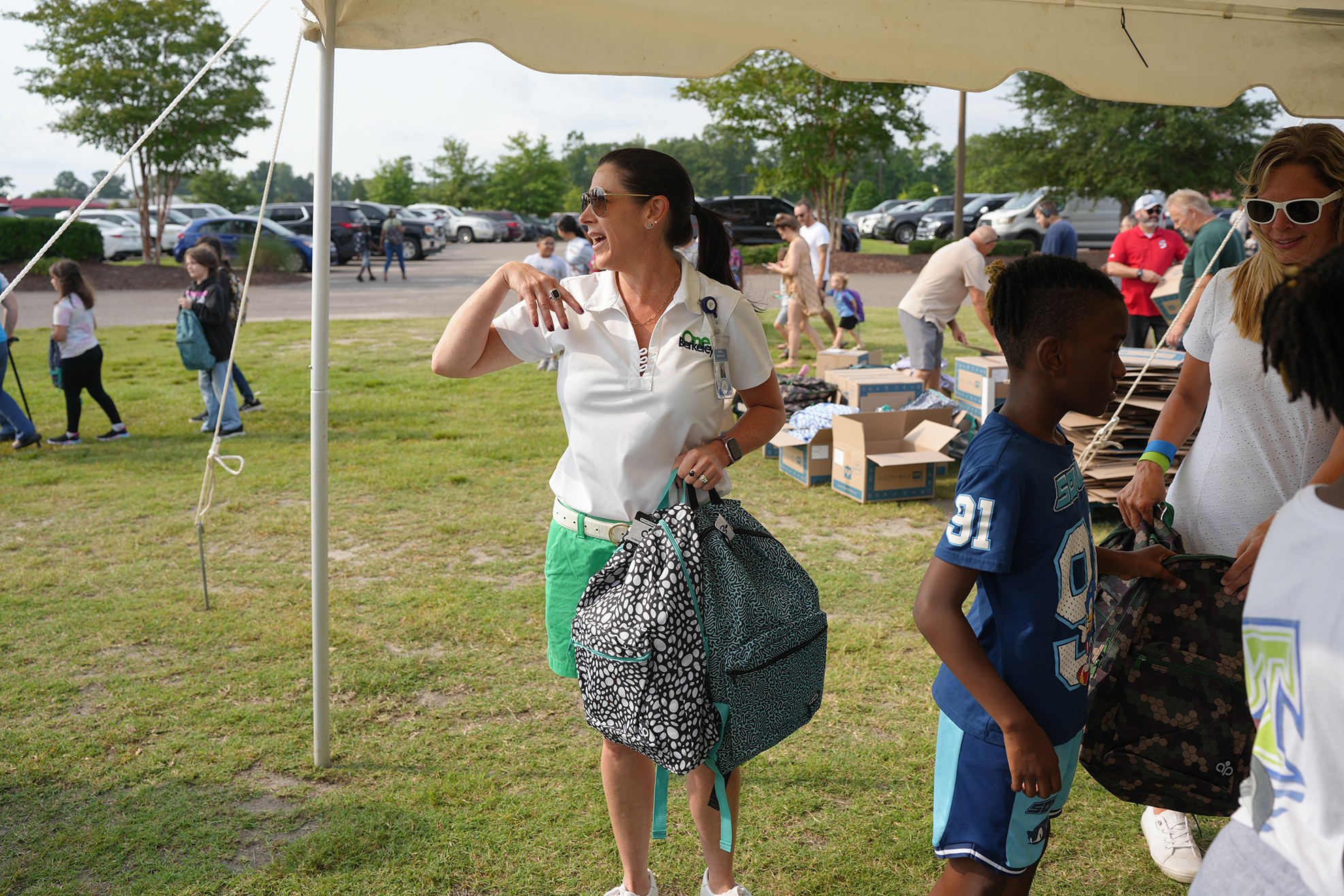 A woman under a tent speaks to a boy with a backpack. People are walking and standing around.