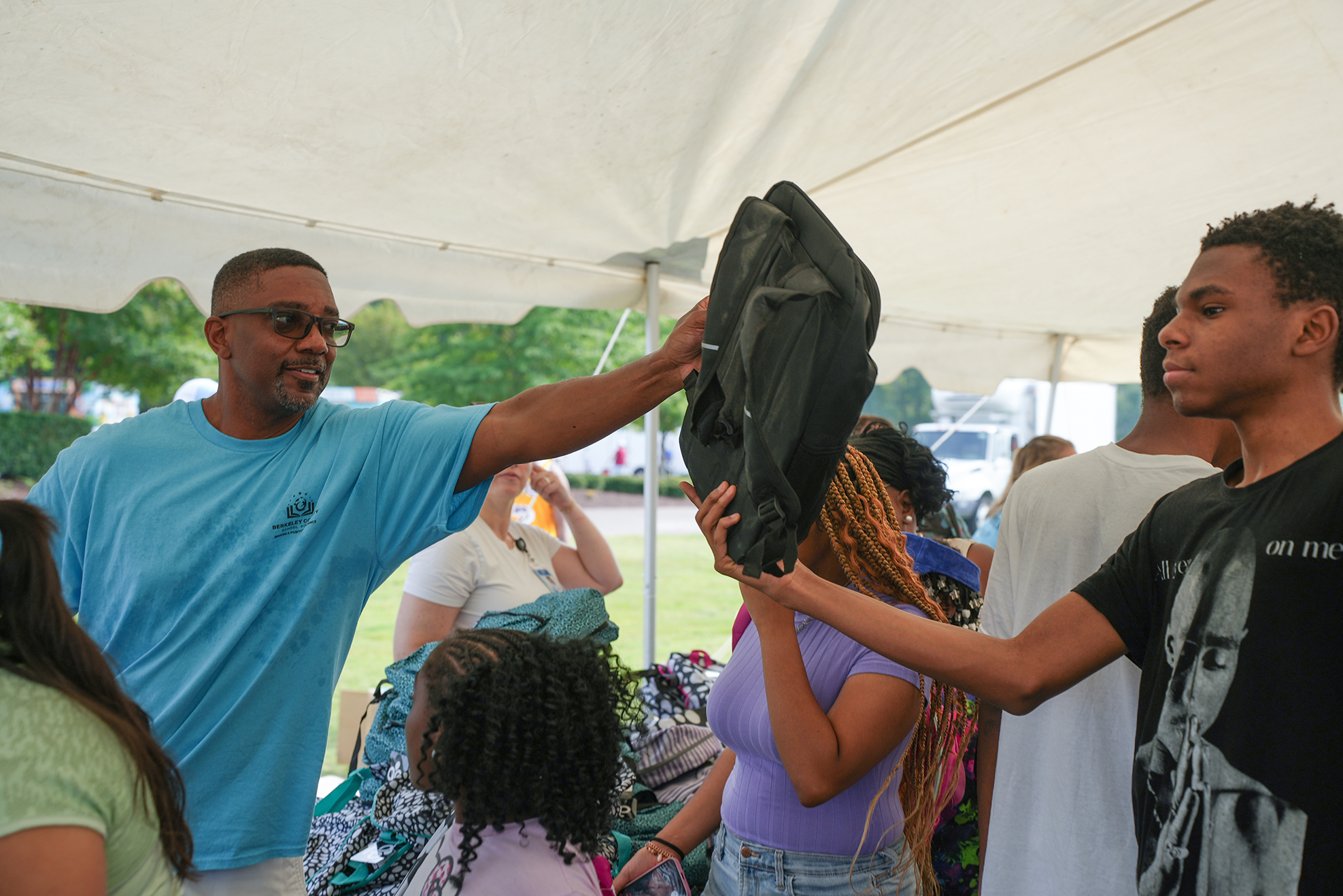 Man in blue shirt and glasses handing jacket to a person holding a black jacket. Others are nearby.