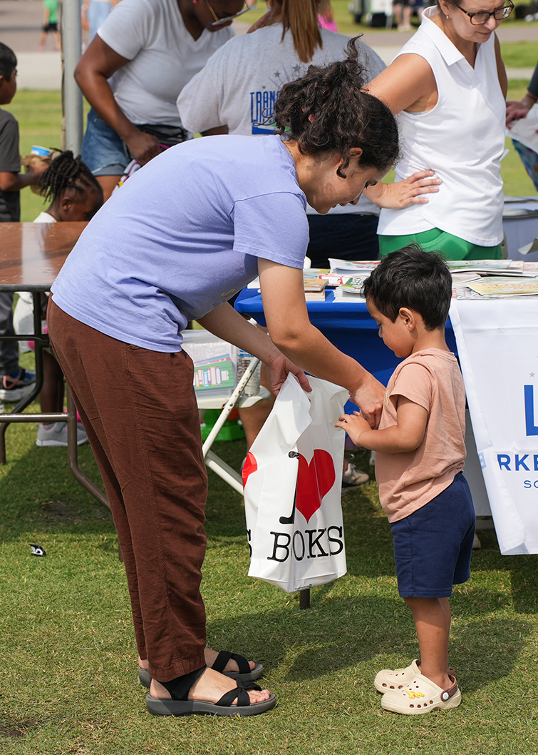 Woman in blue shirt hands a child a white bag with the words "I Heart Books" printed on it.