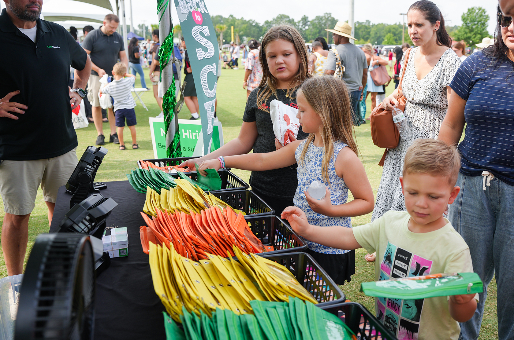 People browse items at a fair or market, with a boy holding a green item and a table full of colorful frisbees.