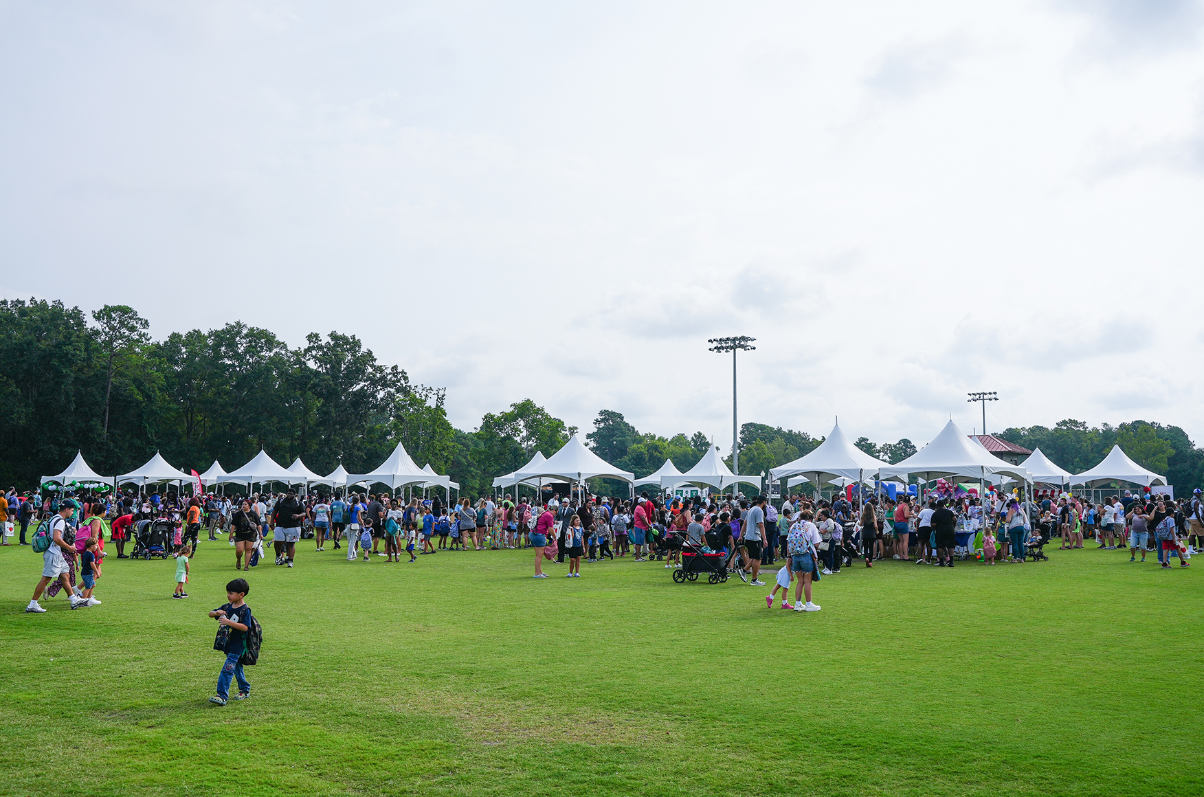 Many people walk around a grassy field. There are white tents in a row. Trees surround the area.