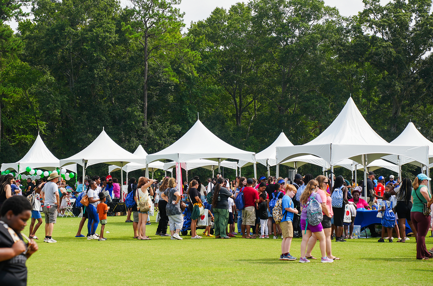 People gather on a grassy field under white tents. Trees are visible in the background.