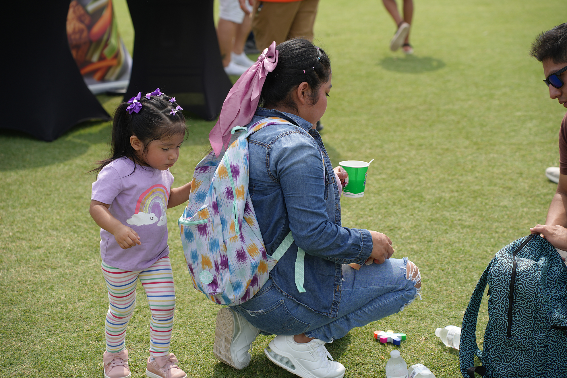 A woman in denim crouches next to a child with a rainbow-themed shirt on a green field.