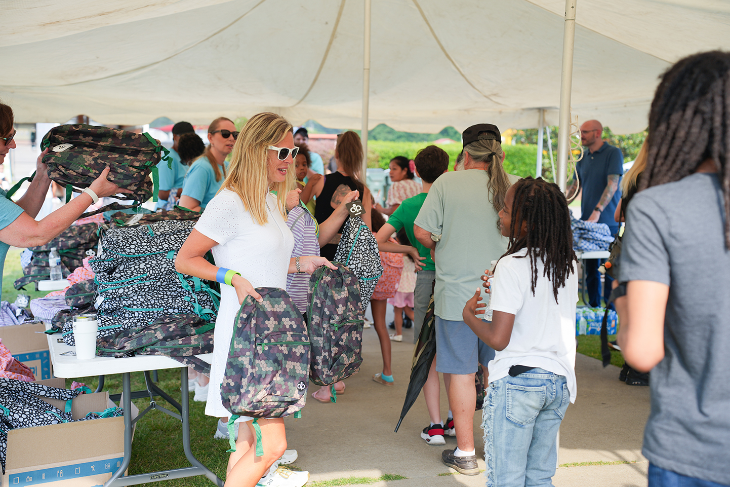 A group of people gather under a tent. A woman holds a backpack. A child holds an umbrella.