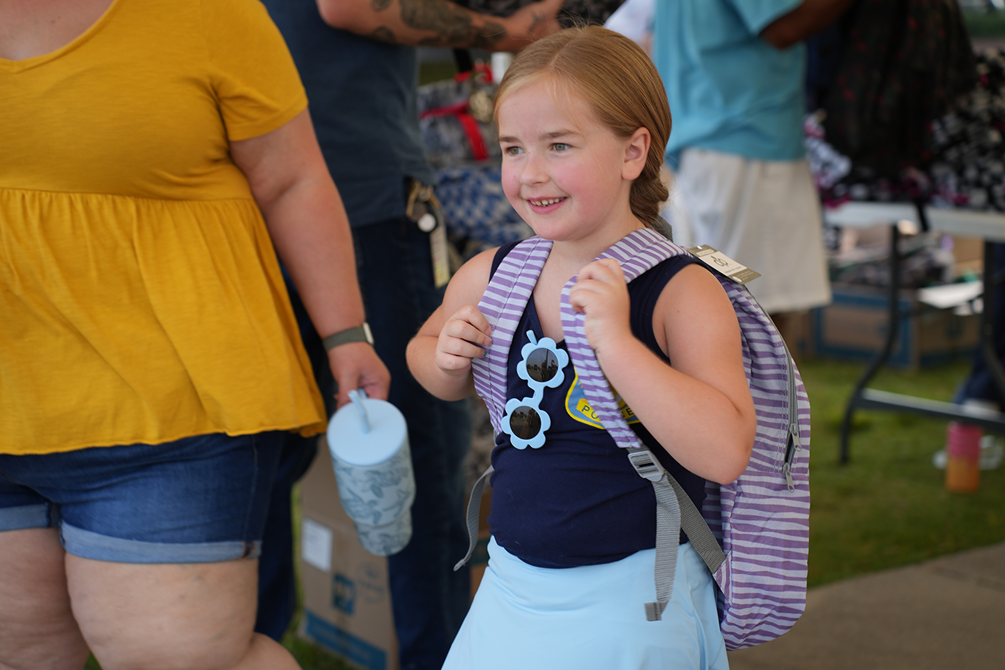 A young girl wearing a backpack, a blue shirt, and a light blue skirt walks outdoors. Behind her, a person in yellow shorts.