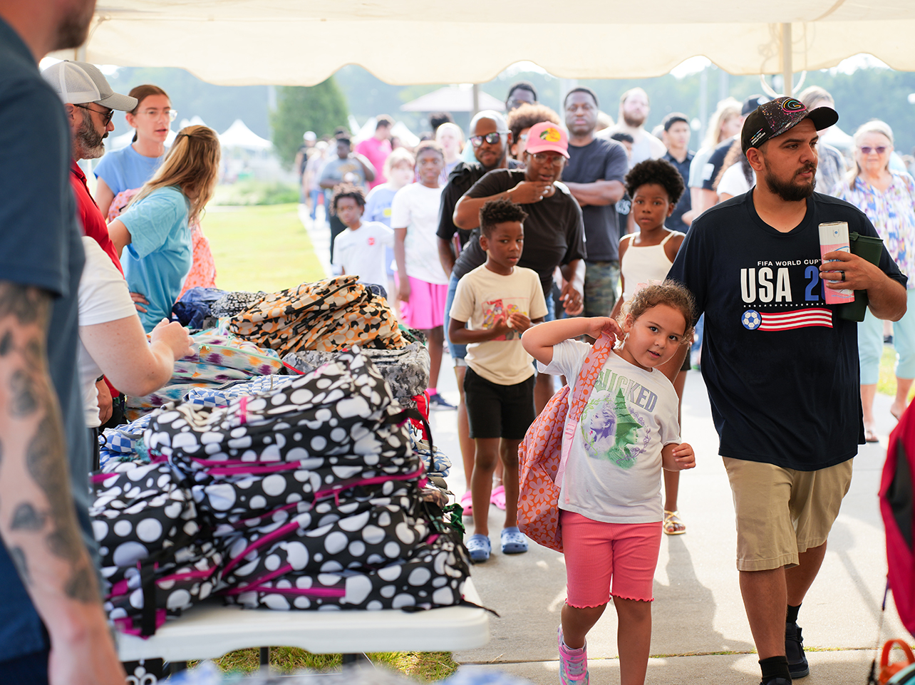 Crowd of people, including a man, woman, and young girl, walking past a table with bags.