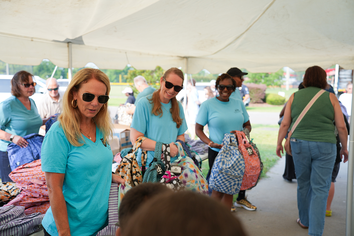 Volunteers in teal shirts stand under a tent, sorting items. Some hold bags, others observe. Background shows a clear sky and greenery.