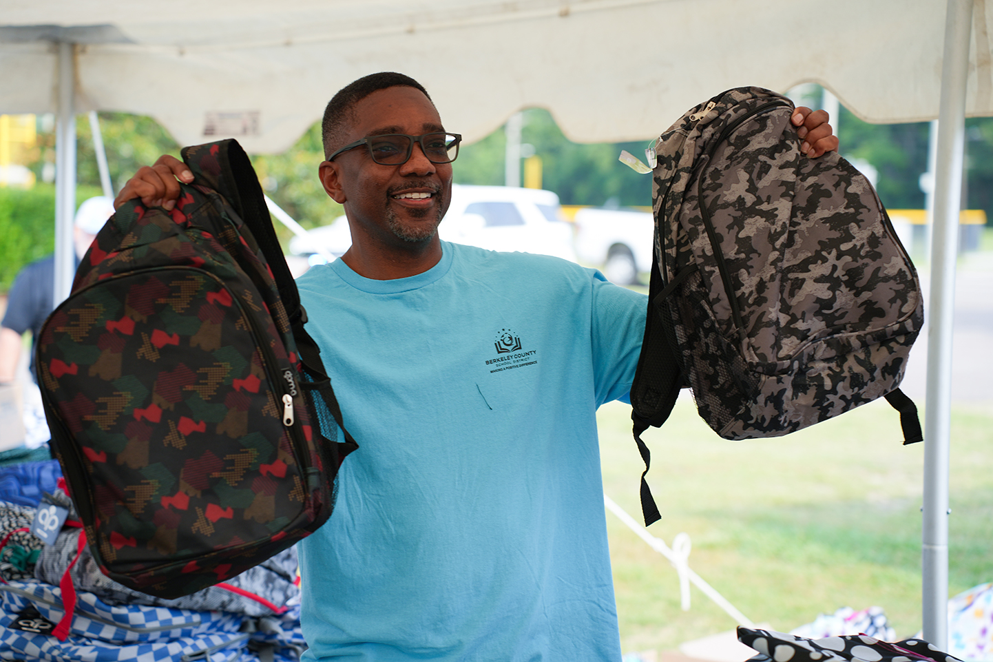 A man in a light blue shirt and glasses holds two camouflage backpacks, standing under a tent.