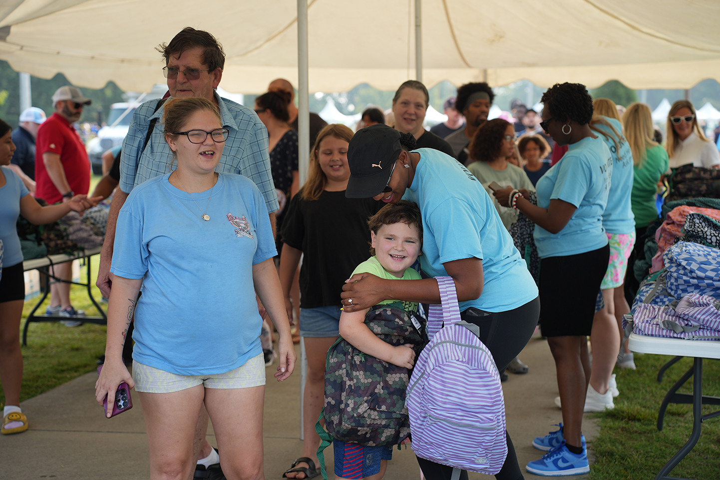 Adults and children stand in a crowd at an outdoor event. Some wear backpacks; others carry bags.
