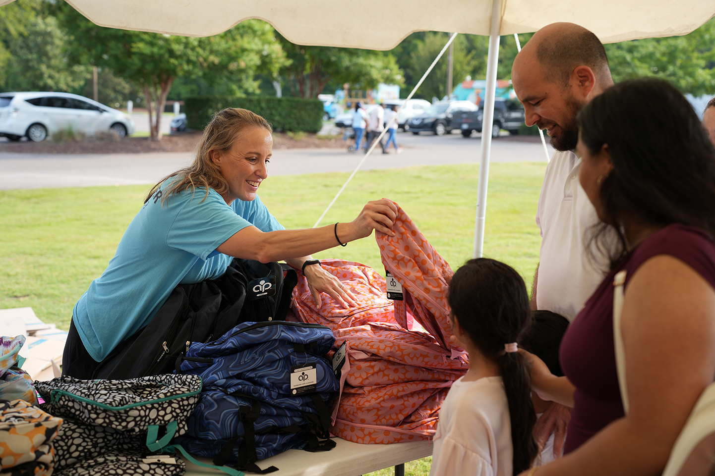 People in a park, one adult holds a child's arm, another smiles, a table holds bags.