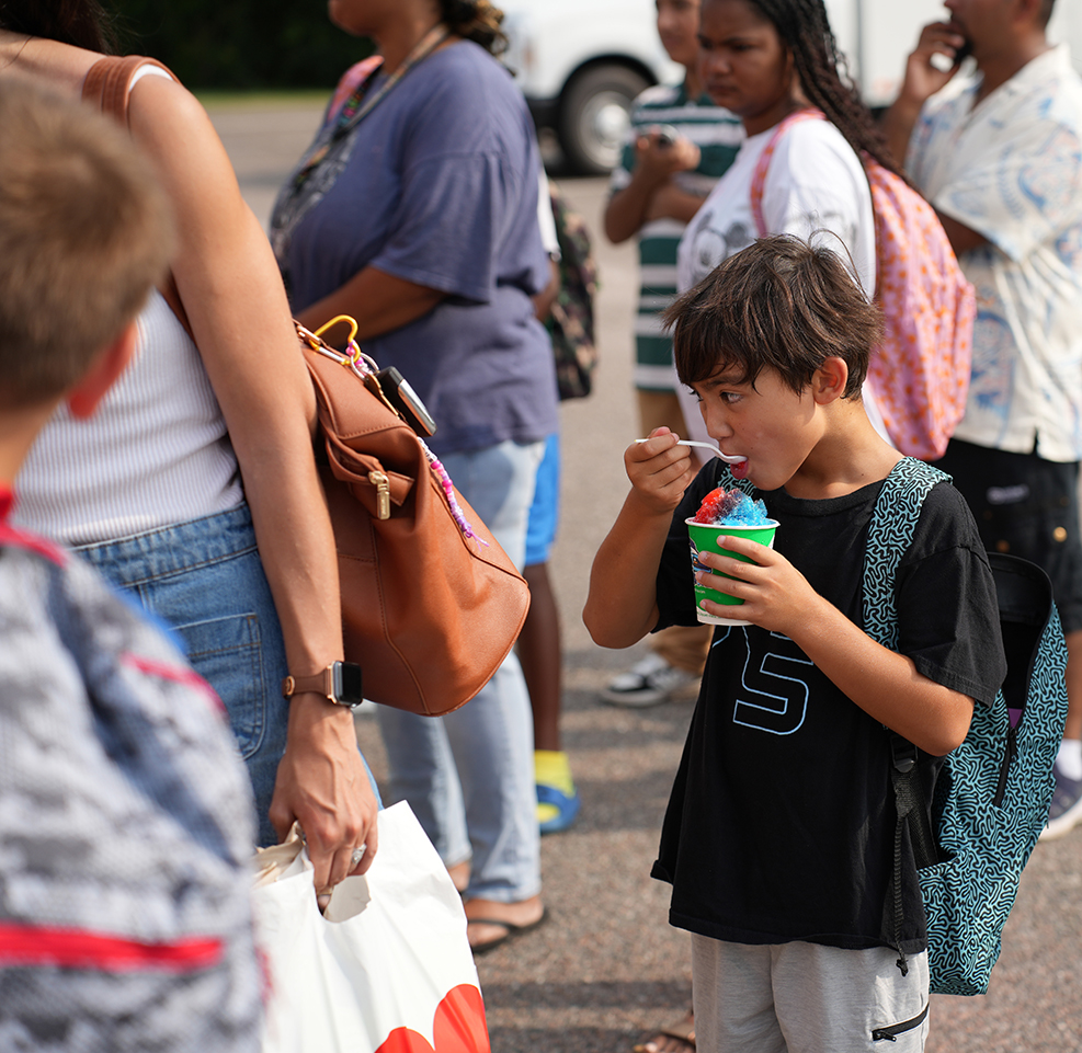 People stand in line outside; one boy eats blue ice cream. Behind him, people hold bags, and a white car is parked.