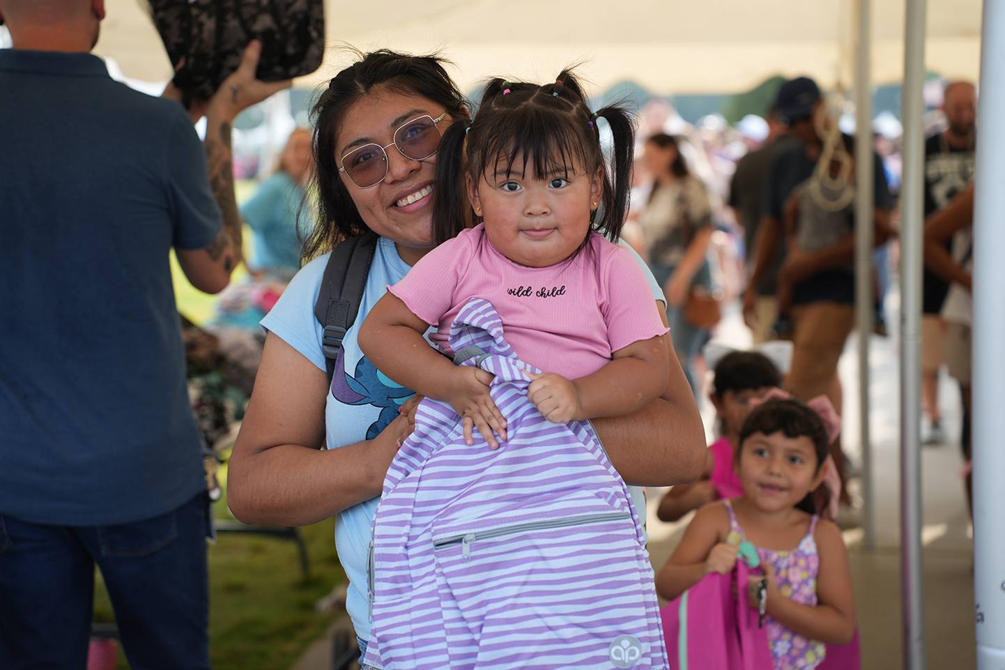 A woman holds a smiling child, both with glasses, at an outdoor event with other people.