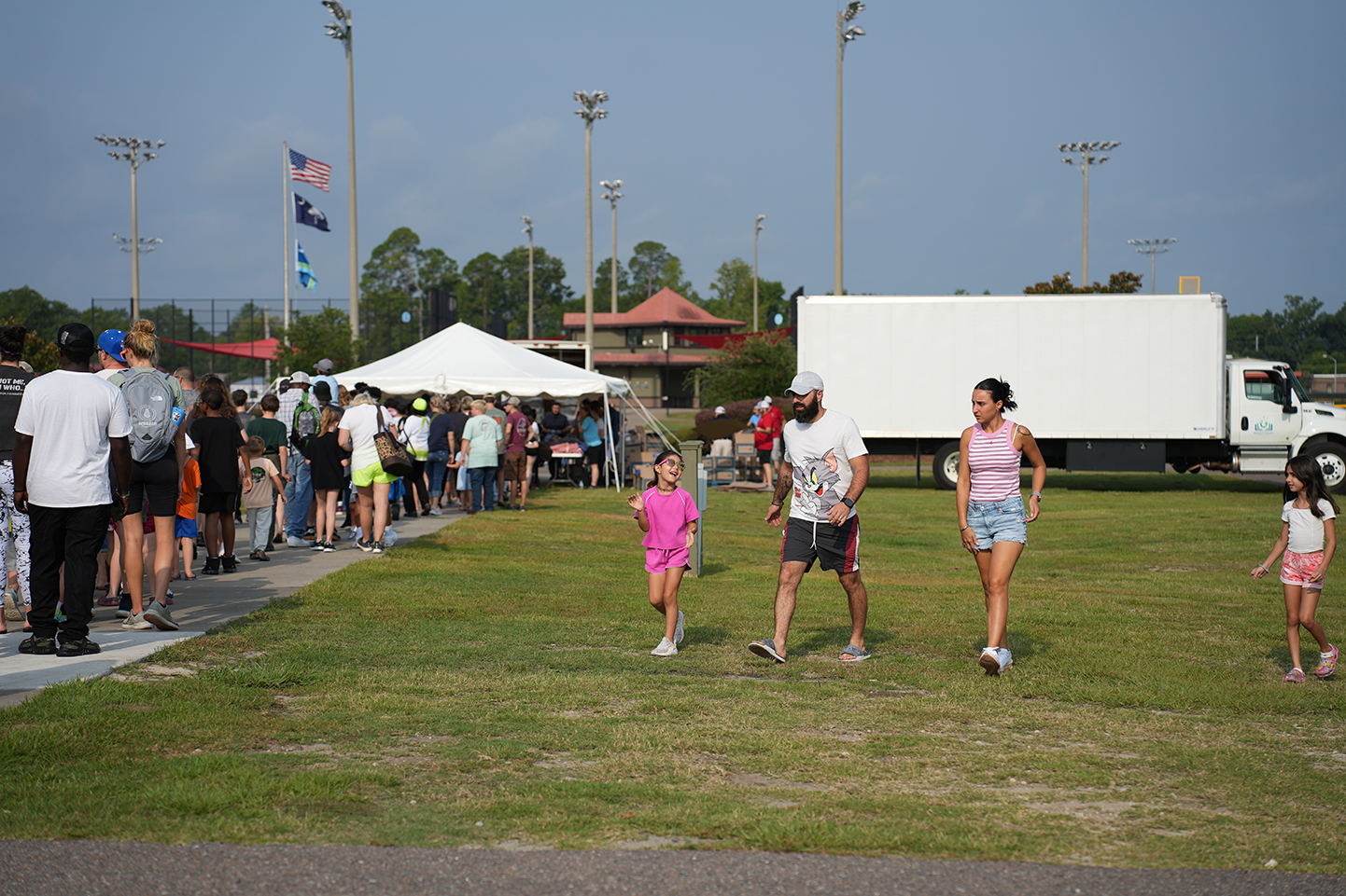 People gather in a field with flags and a white truck. A white tent stands nearby.