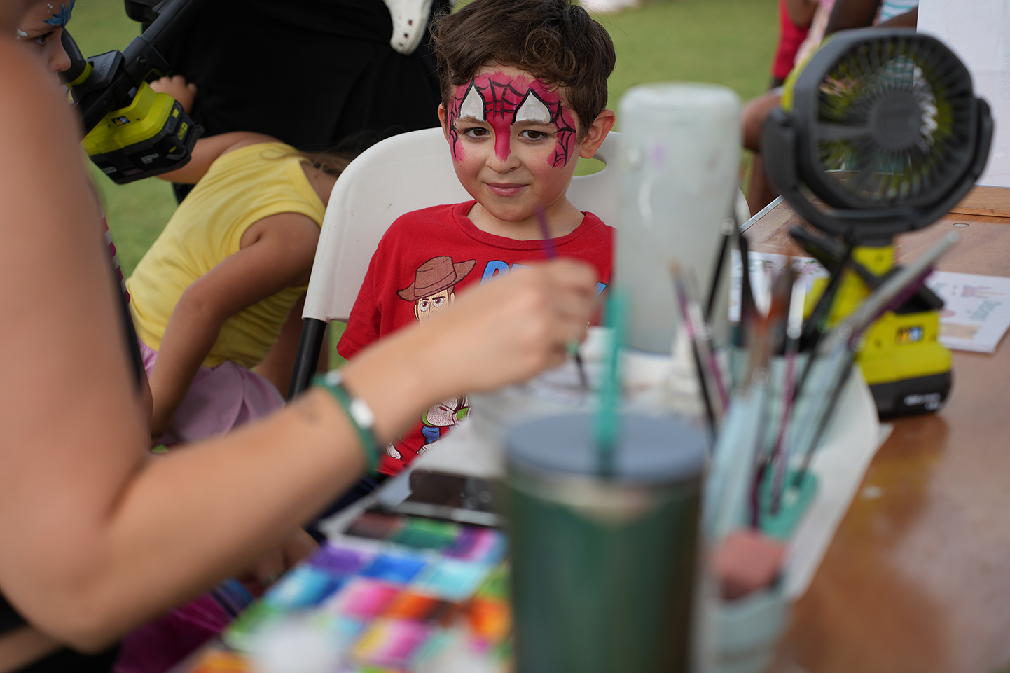 A young boy with spider-man face paint sitting in a chair. A person applies paint on his face.