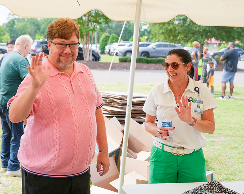 A man in a pink polo waves under a tent. A woman in a white shirt and green pants waves nearby.