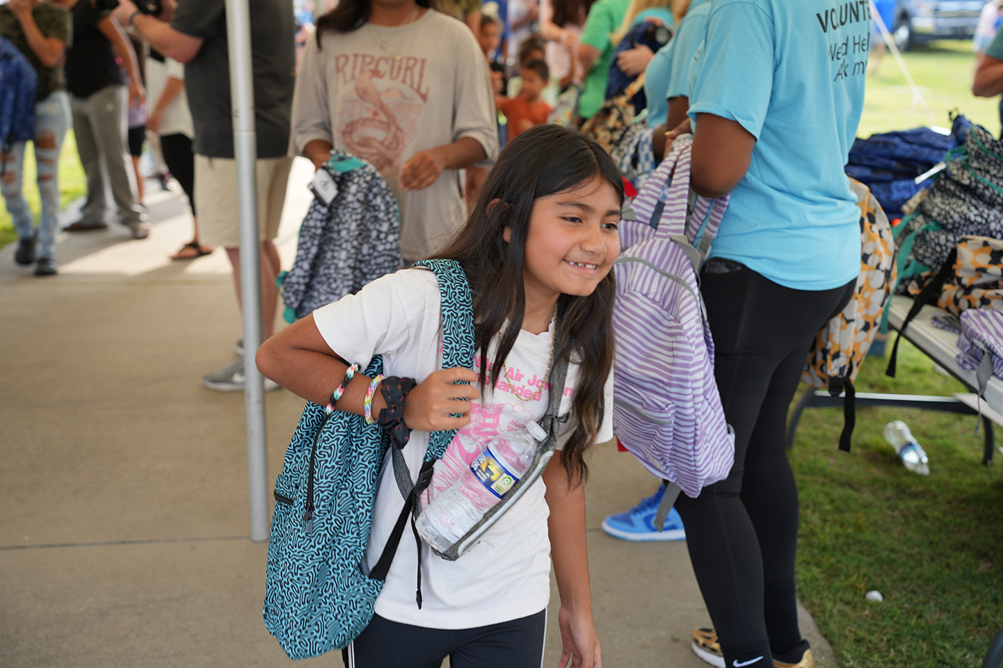 A girl with a backpack, carrying a water bottle, smiles at the camera. Other people and bags are behind her.