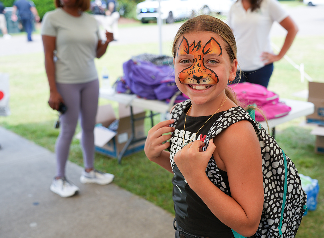 A young girl with a face painted like a tiger, holding a backpack, stands on grass with others.