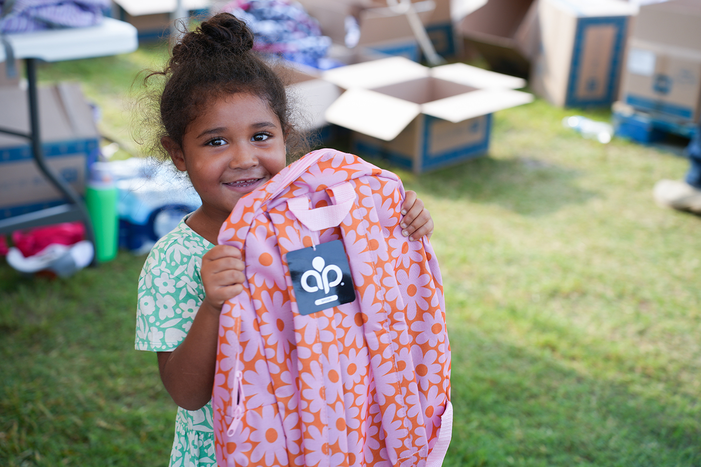 A young girl smiles, holding a pink backpack with an orange pattern and a black tag.