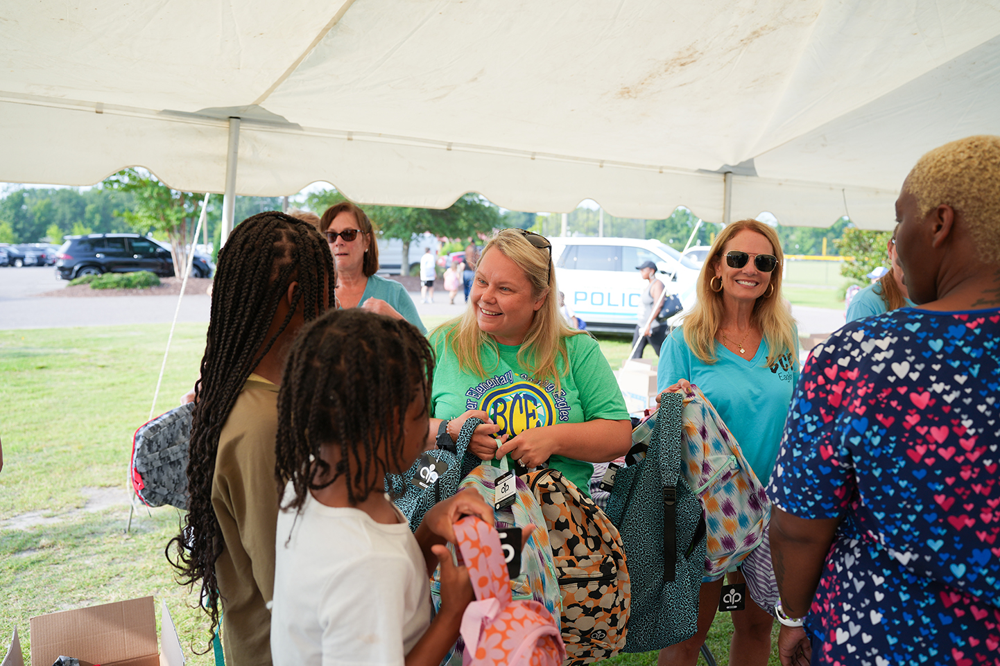 A group of people stands in a white tent. A woman in a green shirt holds bags. A girl holds a pink bag.